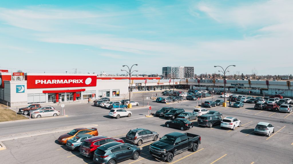 A busy retail parking lot featuring a prominent Pharmaprix store, with various parked cars and a clear blue sky in the background.