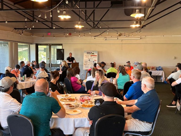 A speaker presents to a large audience at a golf event in a well-lit banquet room, with tables set and attendees engaged, highlighting camaraderie and networking.