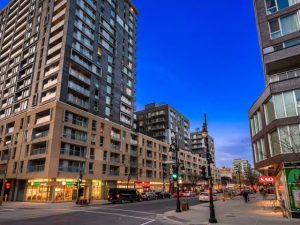 Modern buildings line a bustling street at dusk, with traffic and pedestrians visible. The vibrant blue sky adds to the urban ambiance.