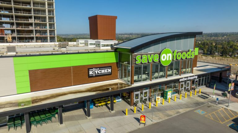 Save-On Foods storefront features vibrant green and brown signage, highlighting the store's kitchen and online shopping options, with construction in the background.