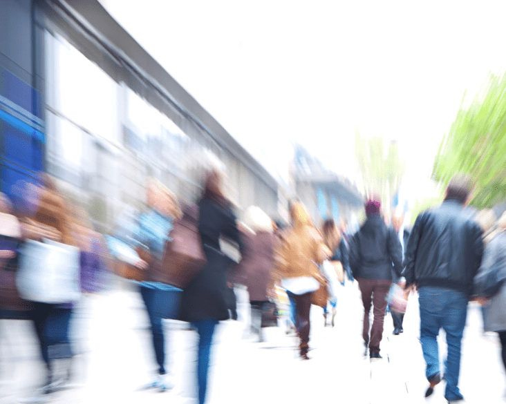 Busy city street filled with blurred pedestrians walking past shops and greenery, illustrating urban life and movement.